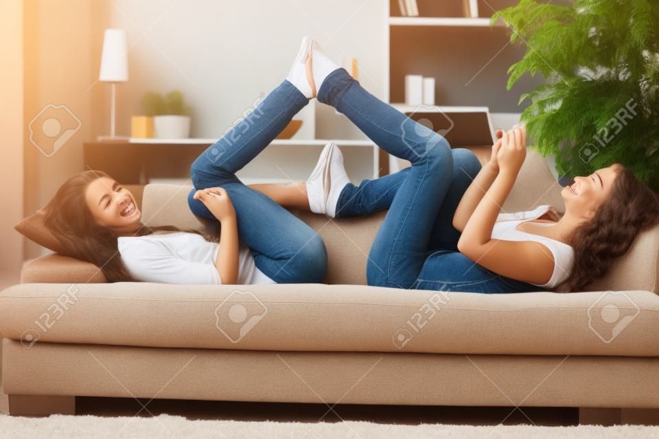 Smiling teens lying on couch with feet put together playing with mobile phone listening to music.�