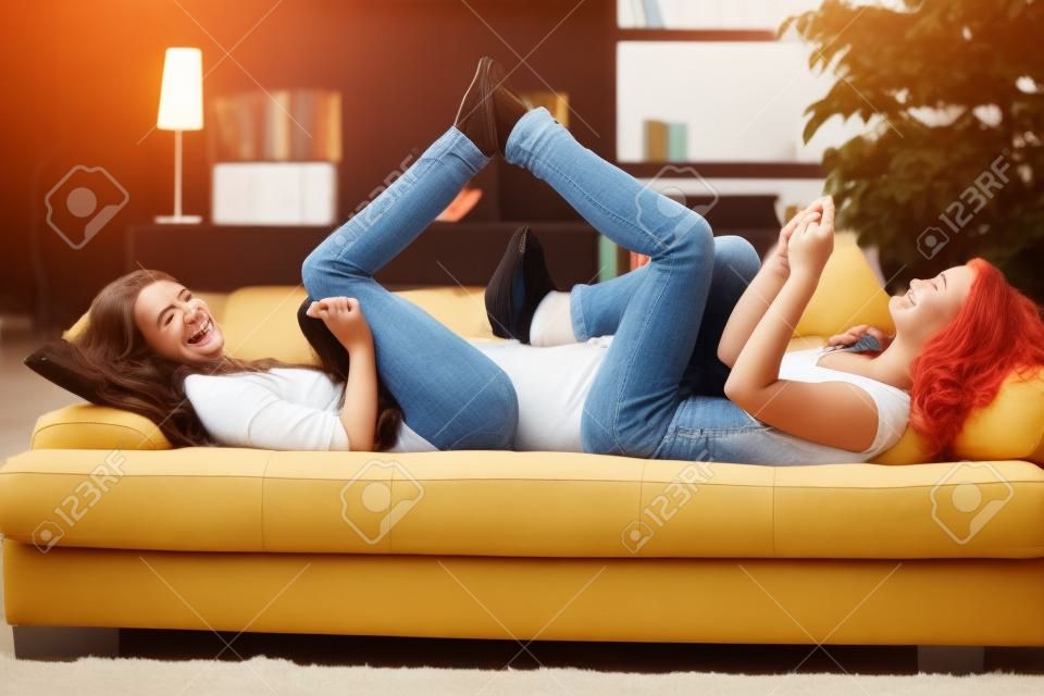 Smiling teens lying on couch with feet put together playing with mobile phone listening to music.�