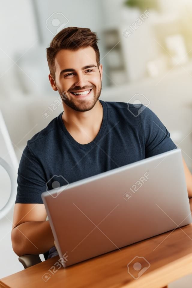 Hombre joven hermoso que usa el ordenador portátil, sonriendo feliz.