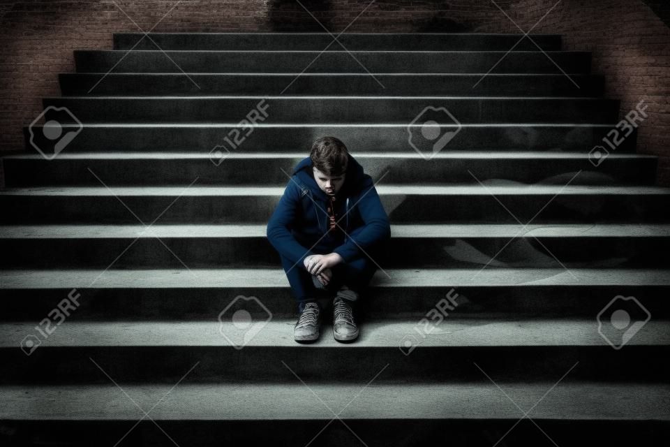 Young desperate man in casual clothes abandoned lost in depression sitting on ground street concrete stairs alone suffering emotional pain, sadness, looking sick in grunge lighting
