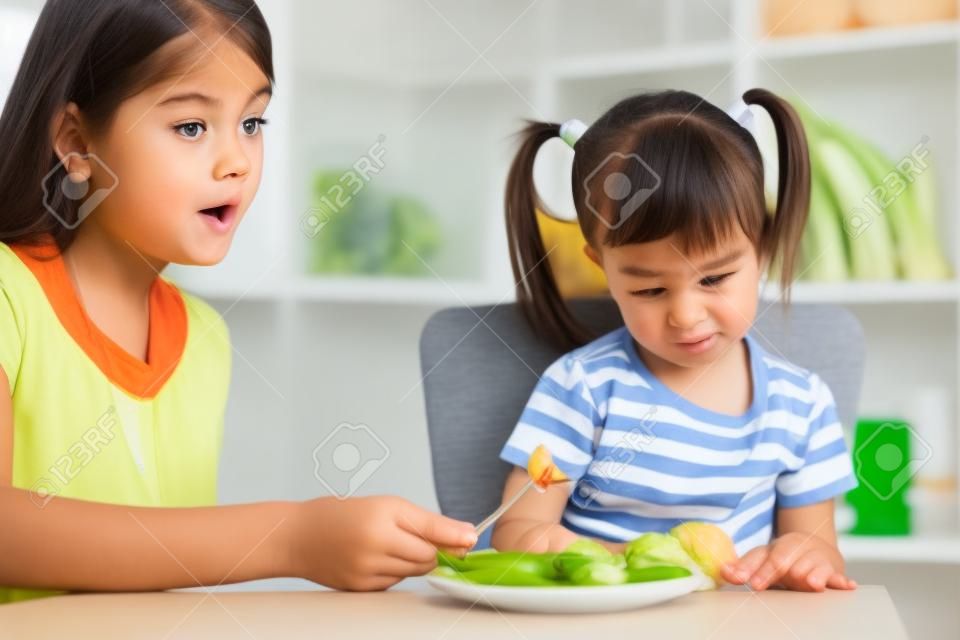 Ребенка девушка смотрит с отвращением на здоровых овощей. Мама уговаривает дочь, чтобы съесть пищу.