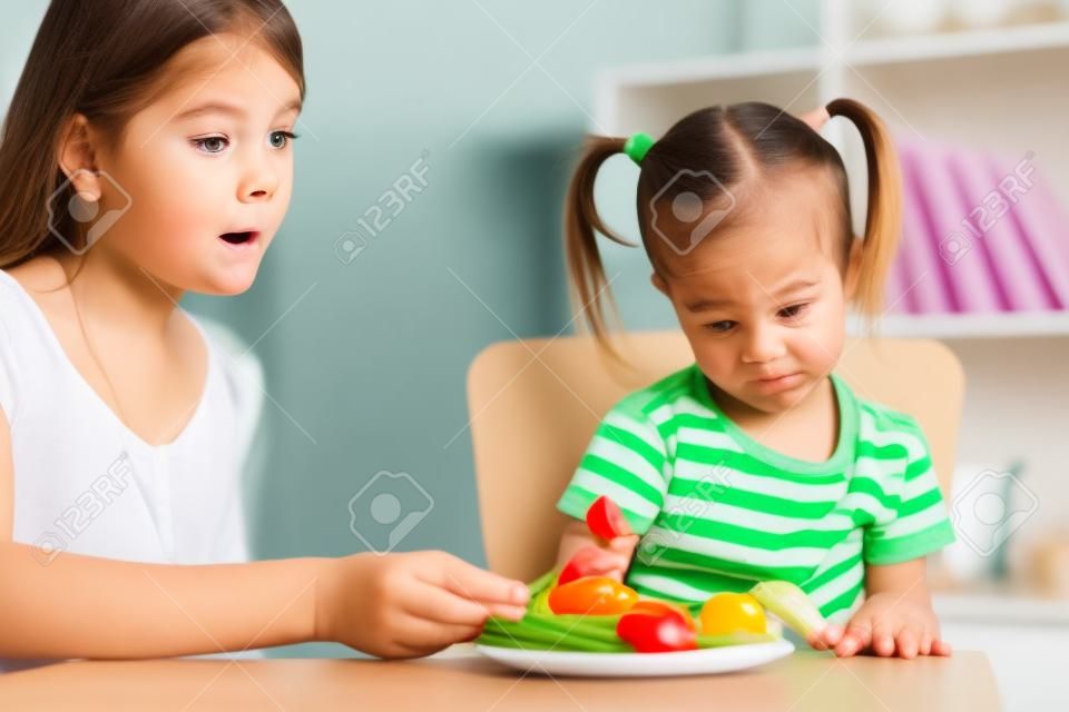 Ребенка девушка смотрит с отвращением на здоровых овощей. Мама уговаривает дочь, чтобы съесть пищу.