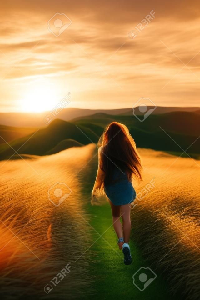 Mulher jovem feliz que corre no campo da grama da pena na luz do por do sol. Estilo de vida da natureza e vista surpreendente ao redor. Cabelo preto longo com fechaduras coloridas