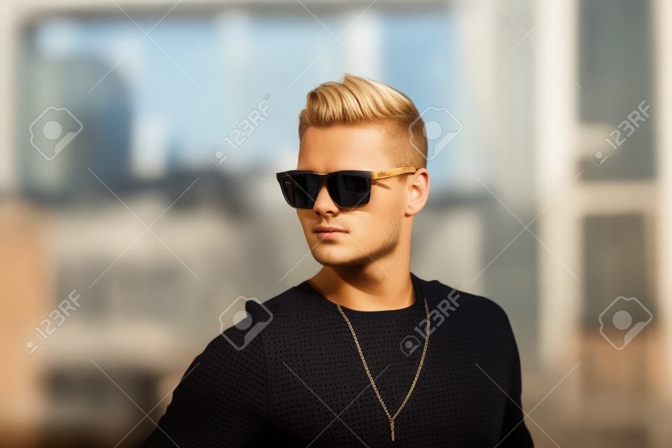 Portrait d'un beau jeune homme aux cheveux blonds courts en lunettes de soleil sur le fond du bâtiment