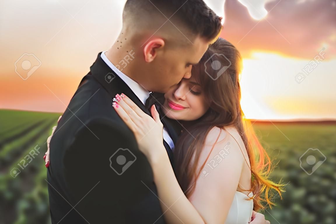 groom is holding and kissing his bride on the background sunset.