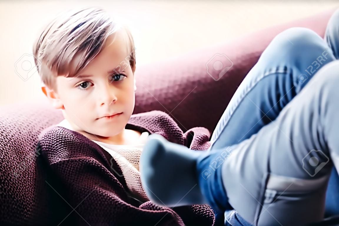 a portrait of a cute boy kid sitting on a sofa against the light window, legs crossed