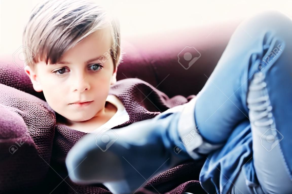 ein Porträt eines niedlichen Jungenkindes, das auf einem Sofa gegen das helle Fenster sitzt, Beine gekreuzt