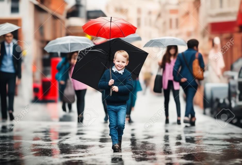 chico lindo con el paraguas caminando en la calle de la ciudad lleno de gente