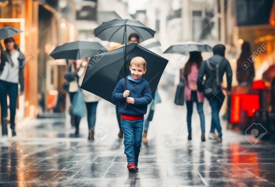 붐비는 도시 거리를 걷고 우산을 가진 귀여운 소년