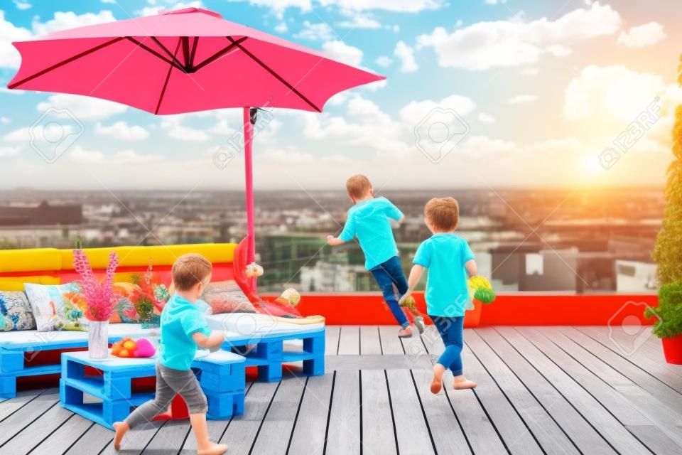 cheerful kids having fun, running on rooftop patio with pallet lounge space, umbrella and decking, home summer activity