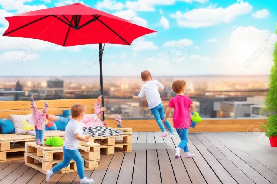 Fröhliche Kinder haben Spaß, laufen auf der Dachterrasse mit Palettenlounge, Sonnenschirm und Terrassendielen, Sommeraktivitäten zu Hause