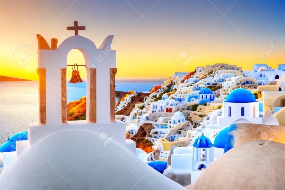 Арка с колоколом, белые дома и церкви с голубыми куполами в Ия или Ia на закате, остров Санторини, Греция