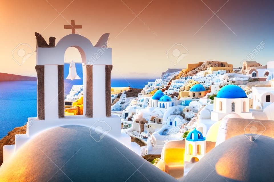 Arch mit einer Glocke, weißen Häusern und Kirche mit blauen Kuppeln in Oia oder Ia bei Sonnenuntergang, Insel Santorini, Griechenland