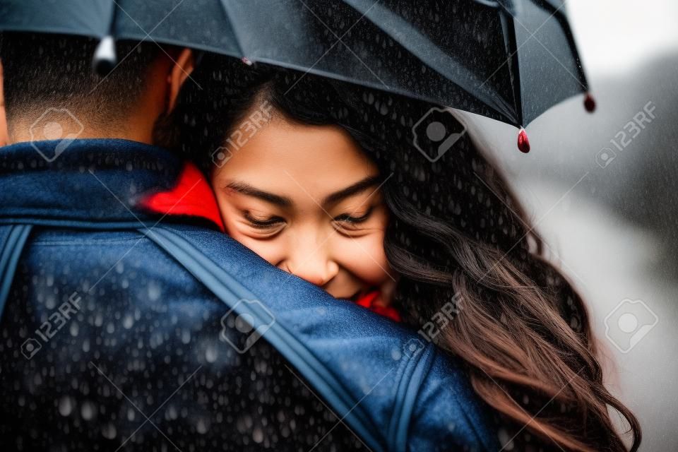 Multiethnic couple of lovers hugging under the umbrella on a rainy day -  Man and woman on a romantic date under the rain, boyfriend hugs his partner to protect her
