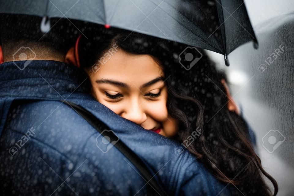 Multiethnic couple of lovers hugging under the umbrella on a rainy day -  Man and woman on a romantic date under the rain, boyfriend hugs his partner to protect her