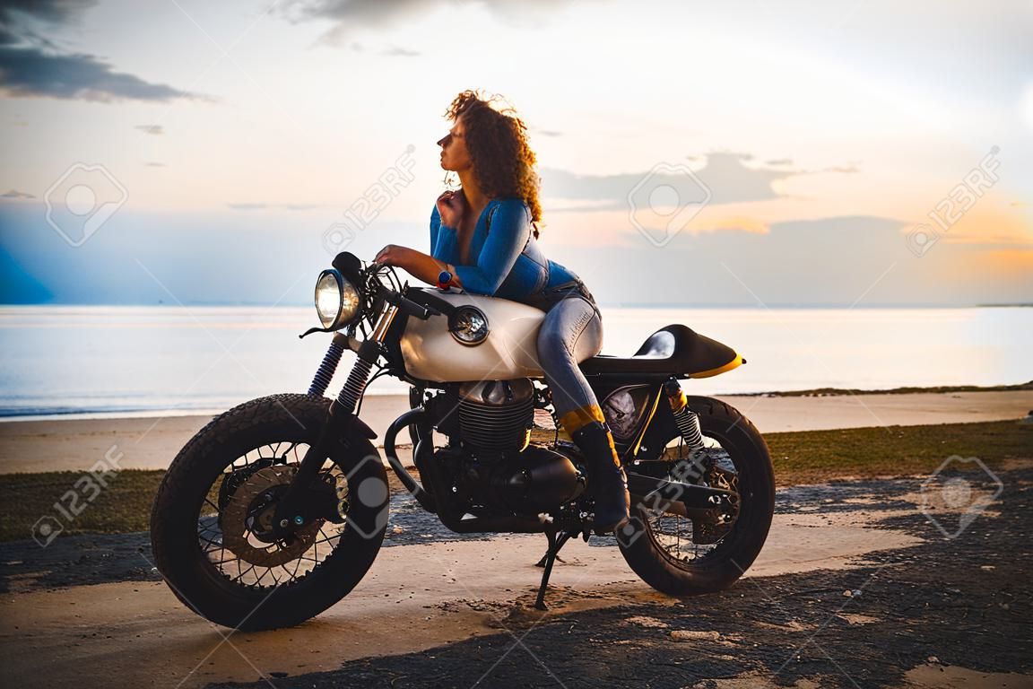 Mooi meisje hebben plezier rijden haar aangepaste cafe racer motorfiets, genieten van de zonsondergang op het strand