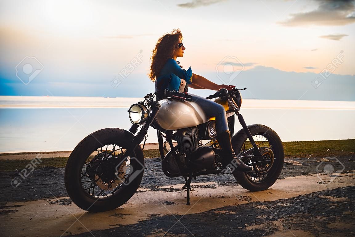Mooi meisje hebben plezier rijden haar aangepaste cafe racer motorfiets, genieten van de zonsondergang op het strand