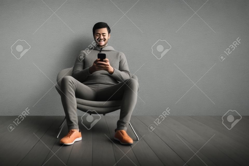 Joyeux gars utilisant un téléphone portable avec une nouvelle application assis dans un fauteuil sur fond de mur gris. gadgets et concept de communication mobile. panorama, espace vide pour le texte
