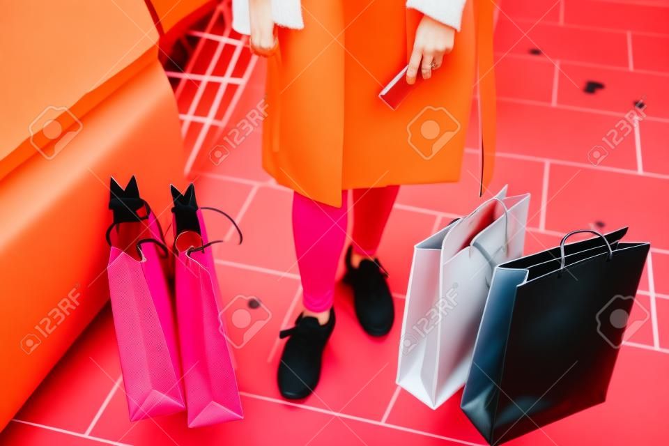Pink, black, orange and white shopping bags resting on the floor next to the feet of a woman wearing jeans