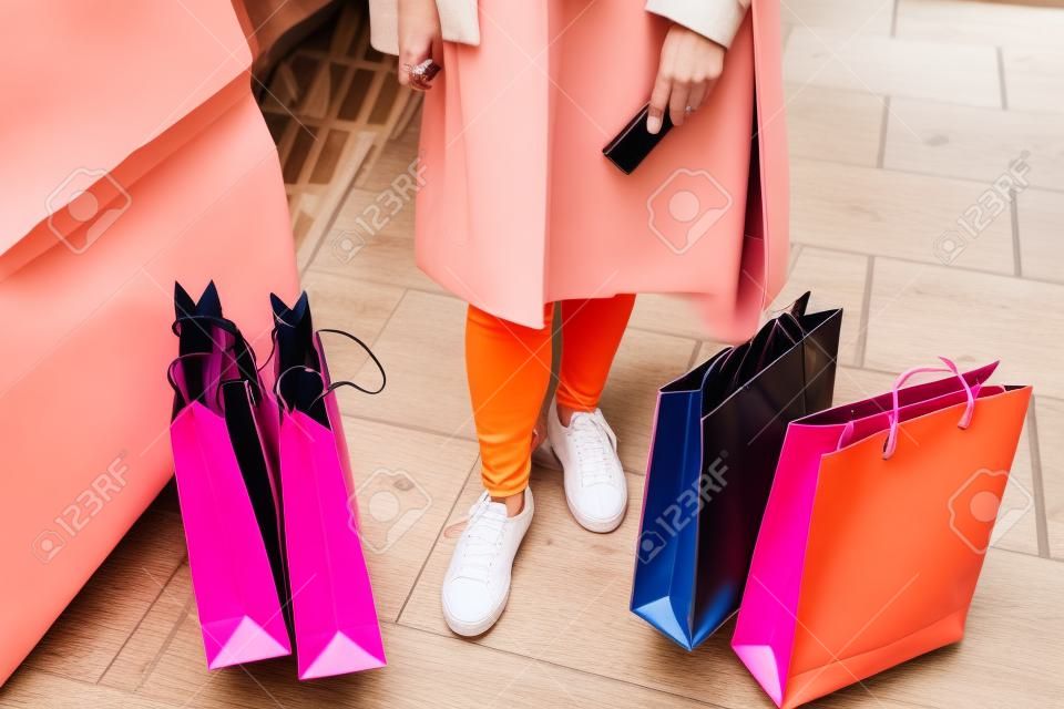 Pink, black, orange and white shopping bags resting on the floor next to the feet of a woman wearing jeans