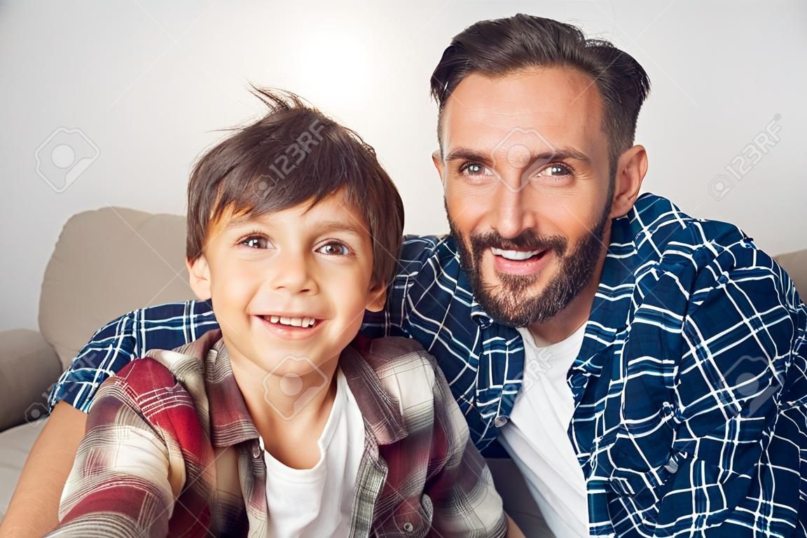 Vater und kleiner Sohn sitzen zu Hause und machen ein Selfie auf dem Smartphone, das die Kamera freudig aus nächster Nähe betrachtet