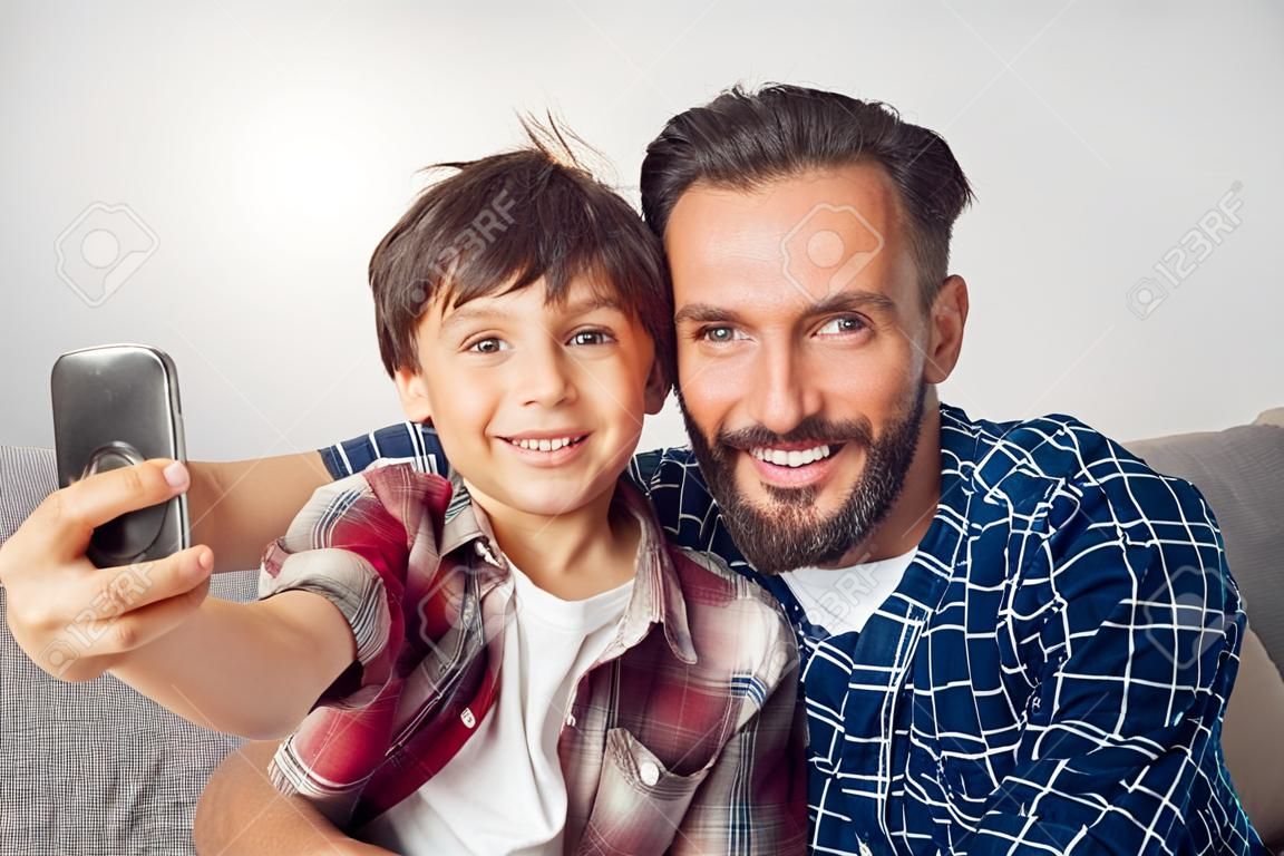 Vater und kleiner Sohn sitzen zu Hause und machen ein Selfie auf dem Smartphone, das die Kamera freudig aus nächster Nähe betrachtet