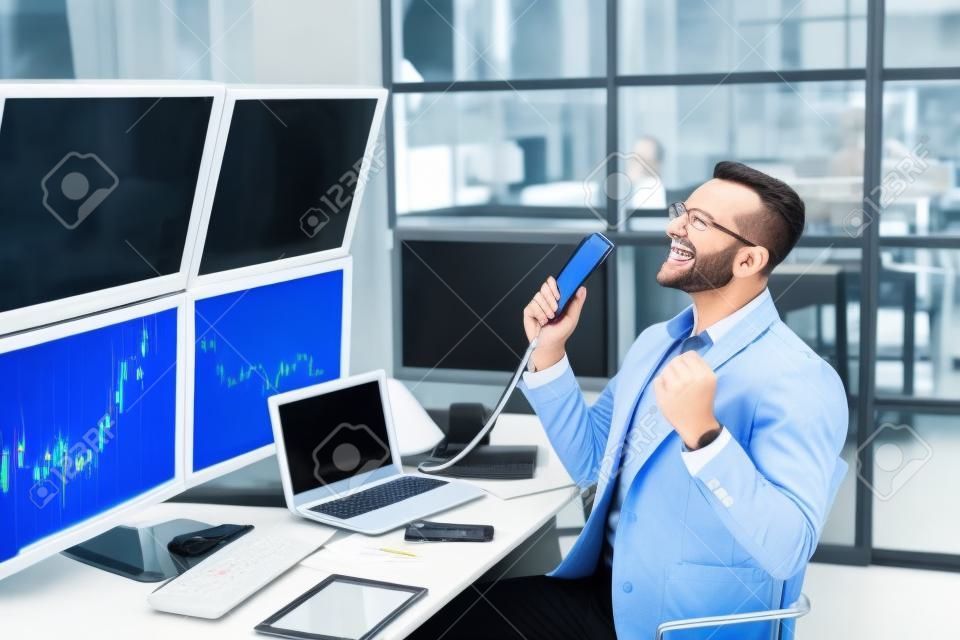 Negociación de acciones. Comerciante sentado en la oficina monitoreando el mercado hablando por teléfono haciendo capacitación comercial en línea riendo alegre en el lugar de trabajo