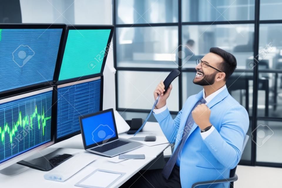 Negociación de acciones. Comerciante sentado en la oficina monitoreando el mercado hablando por teléfono haciendo capacitación comercial en línea riendo alegre en el lugar de trabajo
