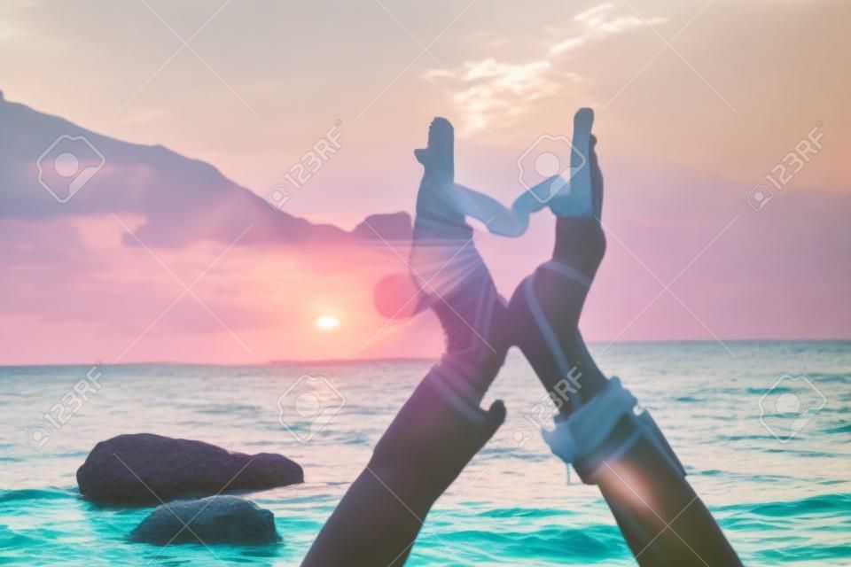 Middle aged well looking woman in white swimsuit and boho style braclets practicing outdoors on yoga mat by the sea on a sunset. Womens yoga fitness routine. Healthy lifestyle, harmony and meditation