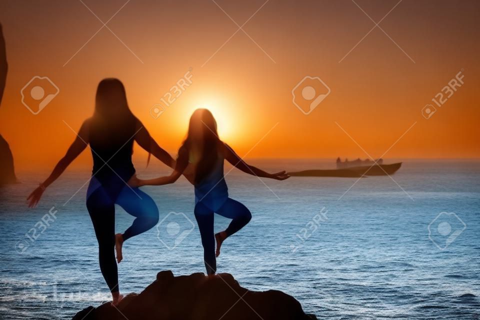 Woman and her daughter practicing balancing yoga pose on one leg up together on rock in the sea. Silhouette mother and daughter doing yoga at beach