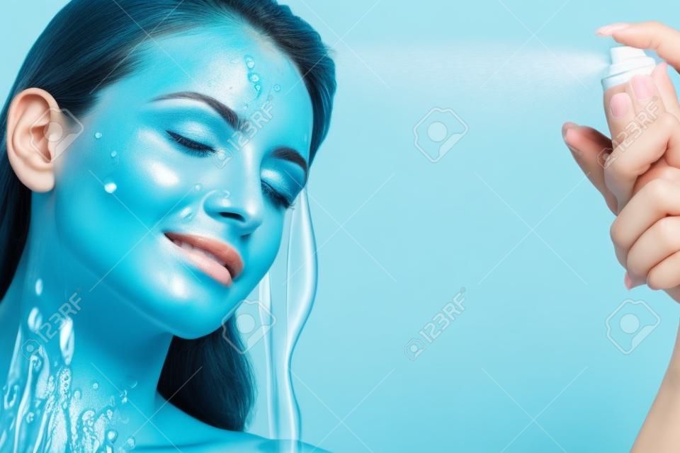 Belle femme appliquant le traitement de l'eau de pulvérisation sur le visage sur le mur bleu