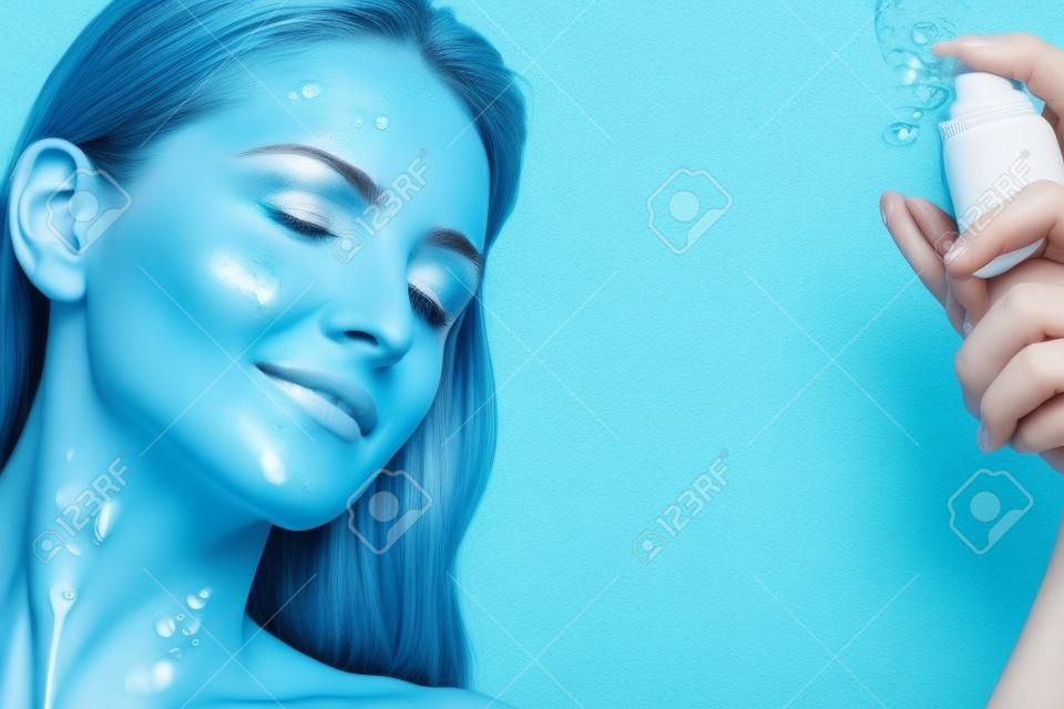 Belle femme appliquant le traitement de l'eau de pulvérisation sur le visage sur le mur bleu