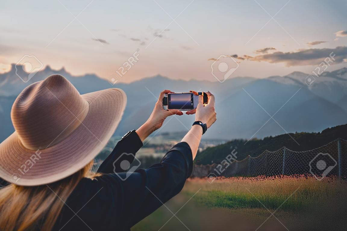 Young woman traveler blogger takes photo on cell phone of Alps mountains at sunset