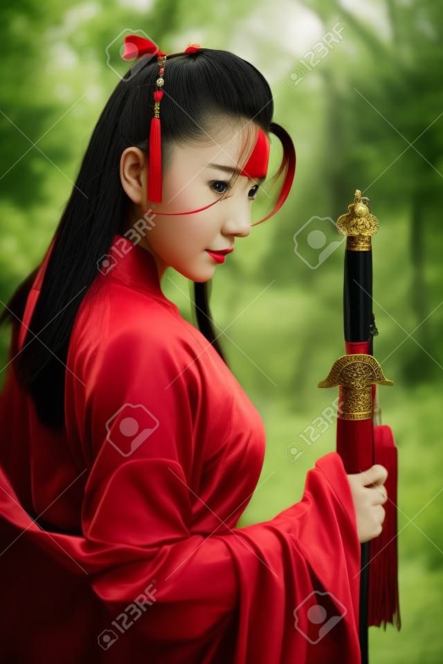 porträt einer jungen und schönen frau, die ein rotes chinesisches kriegerkostüm mit schwarzem schwert trägt, postet sie mit einem schwert zwischen grünen bäumen und der natur im freien