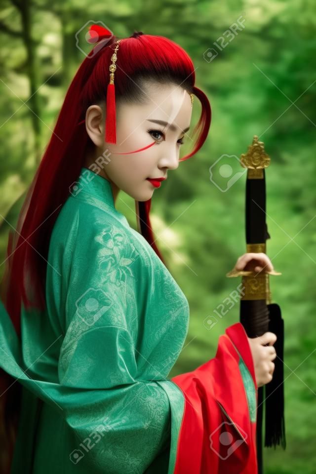 Portret młodej i pięknej kobiety w czerwonym kostiumie chińskiego wojownika z czarnym mieczem, publikuje za pomocą miecza wśród zielonych drzew i przyrody na świeżym powietrzu