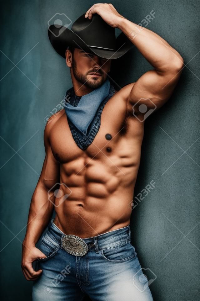 Foto del muscoloso uomo bello che propone in un cappello da cowboy e jeans oltre il muro del grunge.