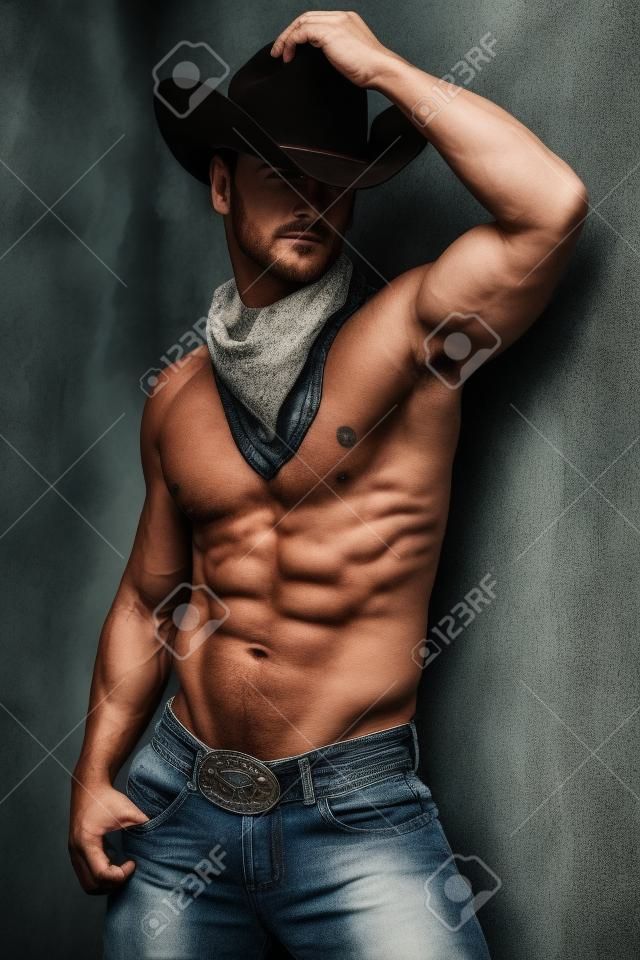 Foto del muscoloso uomo bello che propone in un cappello da cowboy e jeans oltre il muro del grunge.