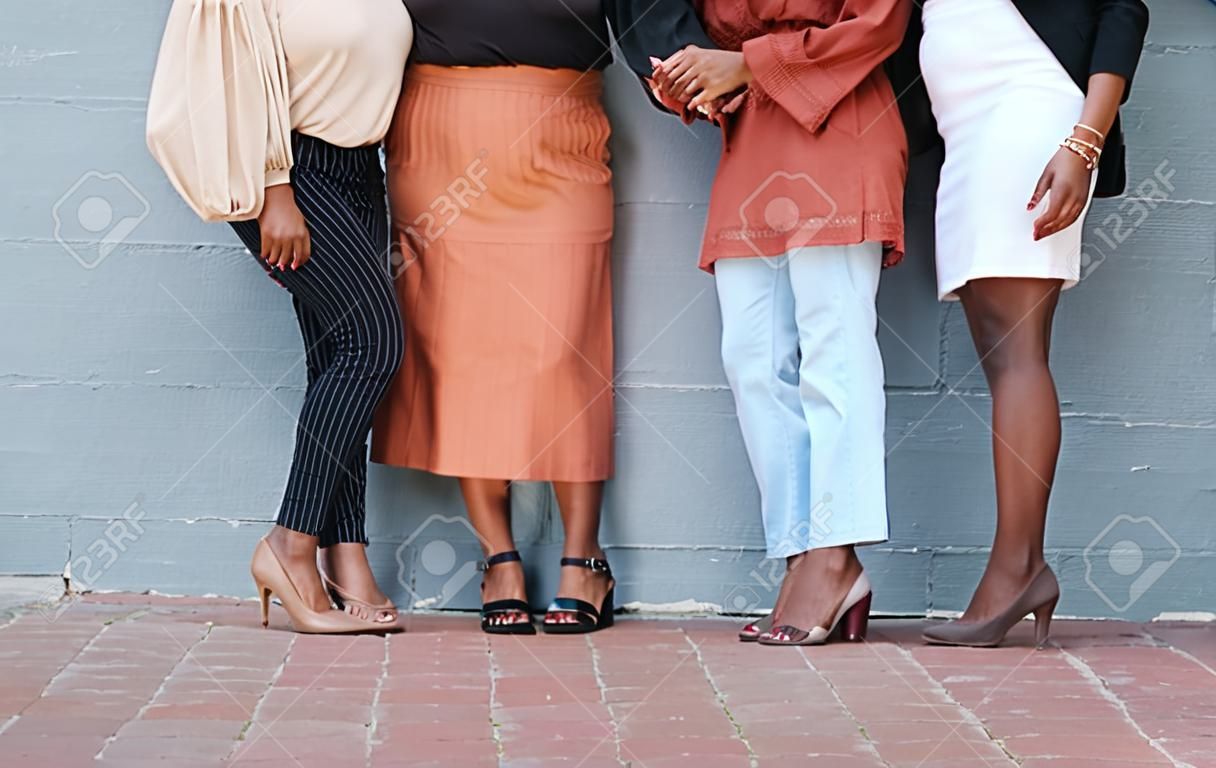 Group of five businesswomen standing against a wall in outside in the city. Legs of friends standing outdoors together from below. Caring businesspeople standing together
