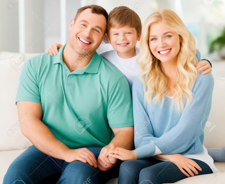 함께 소파에 앉아서 본딩하는 동안 편안하게 보이는 세 명의 행복한 백인 가족. 사랑스러운 작은 금발 소년은 사랑하는 부모와 함께 소파에서 놀면서 그들을 껴안고 있습니다