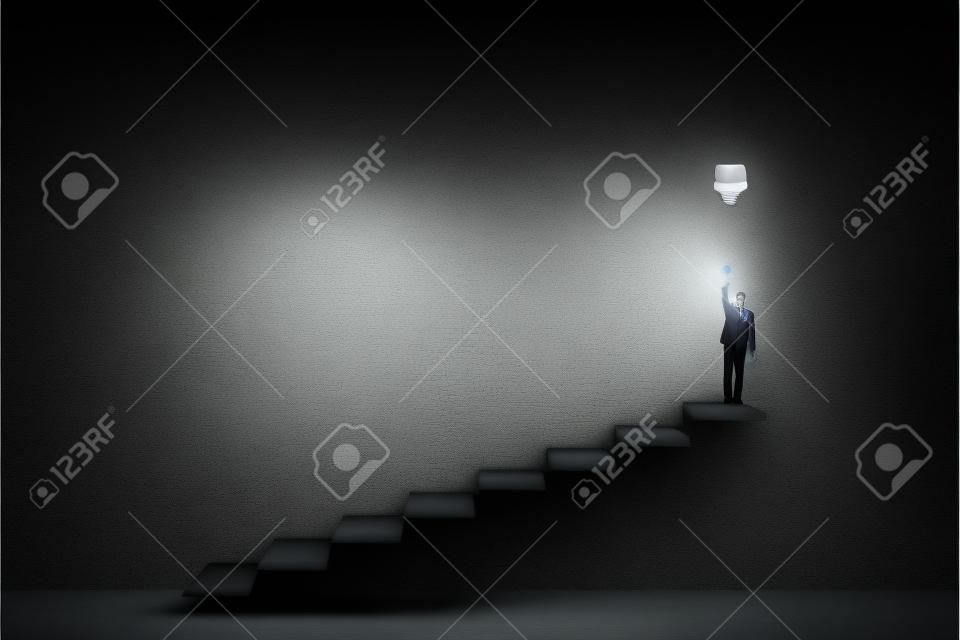 Concepto creativo e idea con vista frontal sobre el hombre de traje en la parte superior de la escalera encendiendo una gran bombilla sobre fondo de hormigón oscuro en una sala vacía