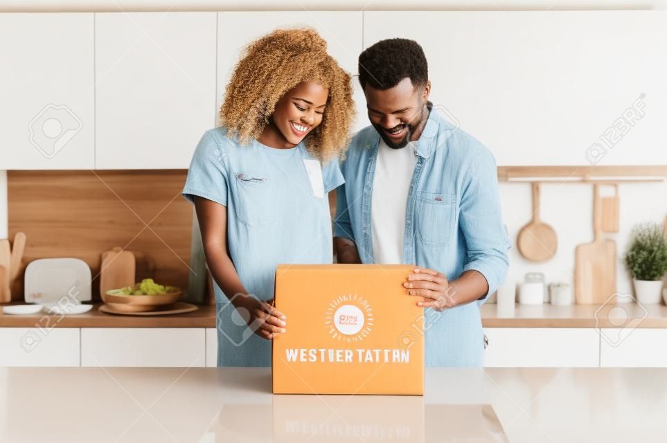 Happy smiling interracial couple standing by the table in the modern kitchen and opening a carton box, young millennial family excited to receive their wedding or housewarming gift, unboxing concept