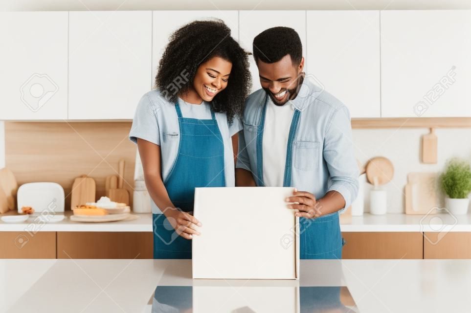 Casal interracial sorridente feliz que está ao lado da mesa na cozinha moderna e que abre uma caixa da caixa, família jovem millennial excitada para receber seu presente do casamento ou do housewarming, conceito do unboxing