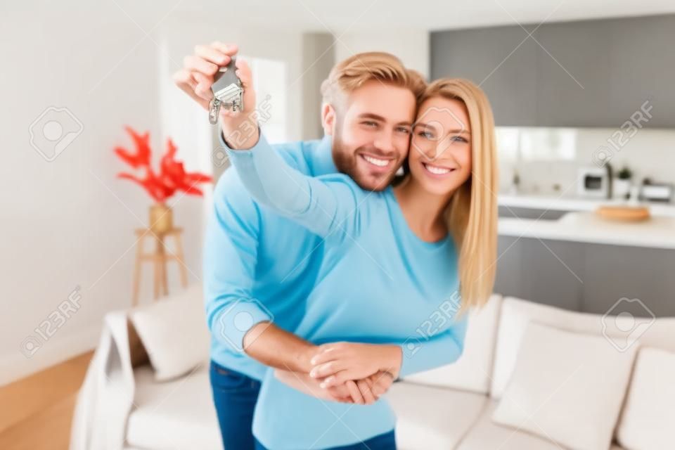 Feliz pareja de familia caucásica mostrando nuevas llaves de casa a la cámara mientras posa en el interior. Vivienda propia, propiedad inmobiliaria y vivienda. Hipoteca y compra de apartamento.
