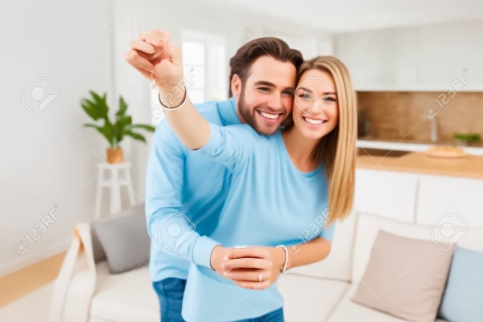 Feliz pareja de familia caucásica mostrando nuevas llaves de casa a la cámara mientras posa en el interior. Vivienda propia, propiedad inmobiliaria y vivienda. Hipoteca y compra de apartamento.