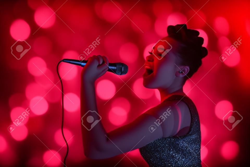 Mulher adolescente bonita que canta o artista do concerto do kareoke que guarda o microfone, no fundo de luzes desfocadas laranja vermelha.