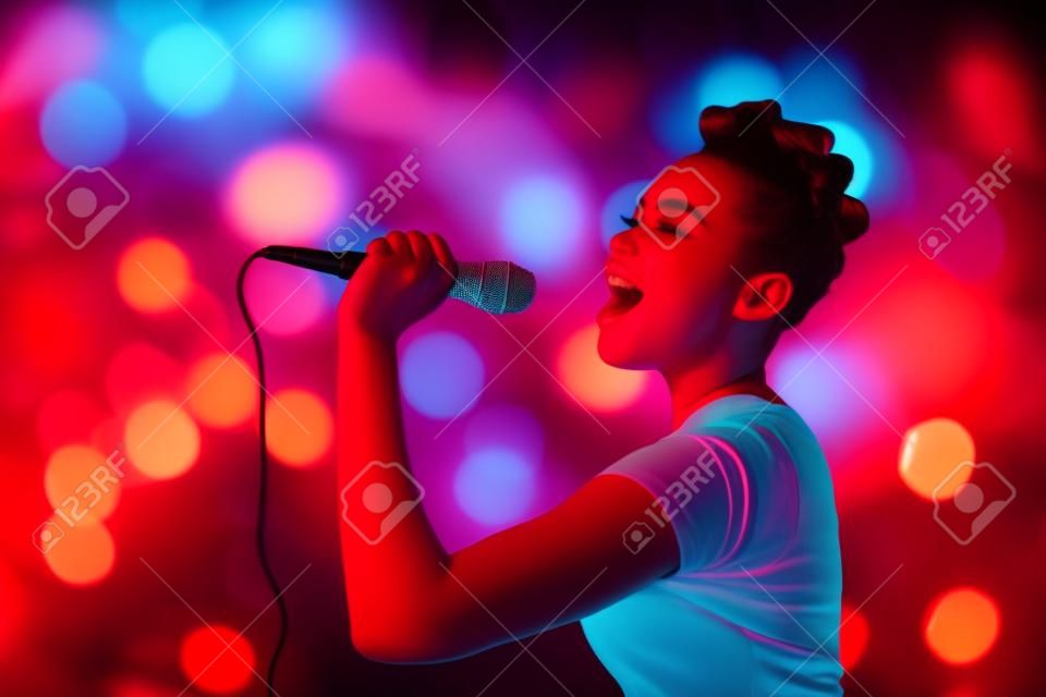 Красивая подросток женщина пение караоке концерт художник держит микрофон, на красный оранжевый размытым фоном огни.