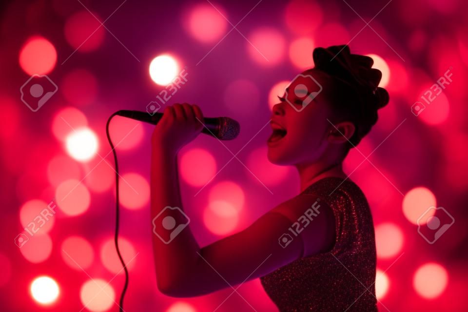 Hermosa mujer adolescente cantando artista de concierto de kareoke con micrófono, sobre fondo rojo naranja borrosa luces.