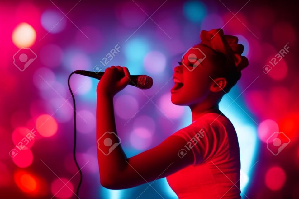 Mulher adolescente bonita que canta o artista do concerto do kareoke que guarda o microfone, no fundo de luzes desfocadas laranja vermelha.