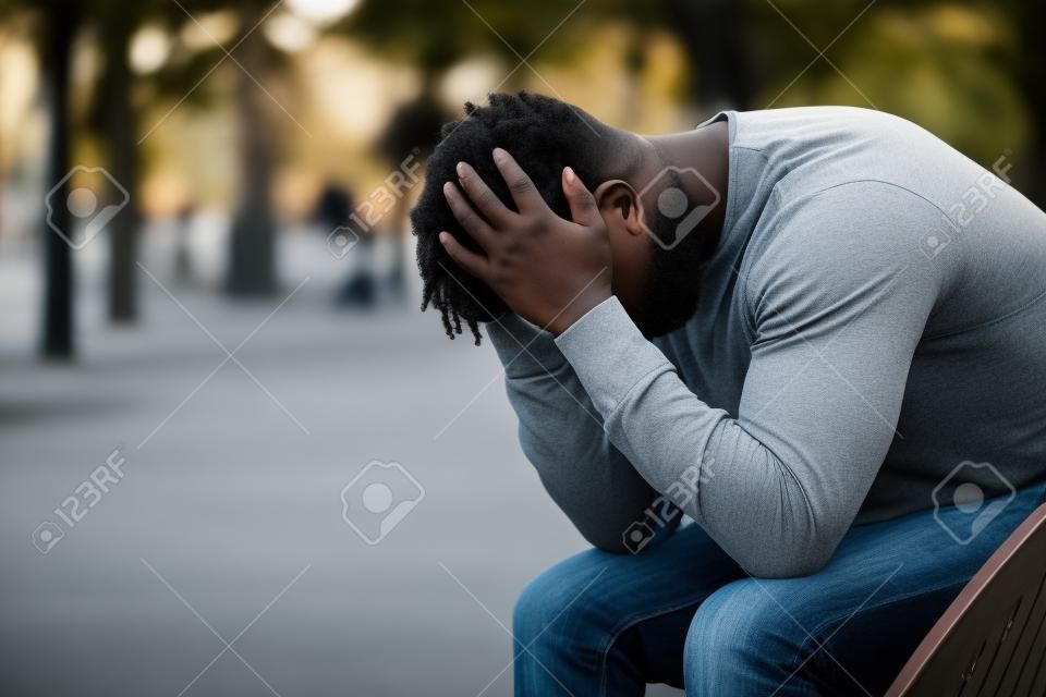 Seitenansichtporträt eines traurigen depressiven schwarzen Mannes, der auf einer Bank in einem Park sitzt