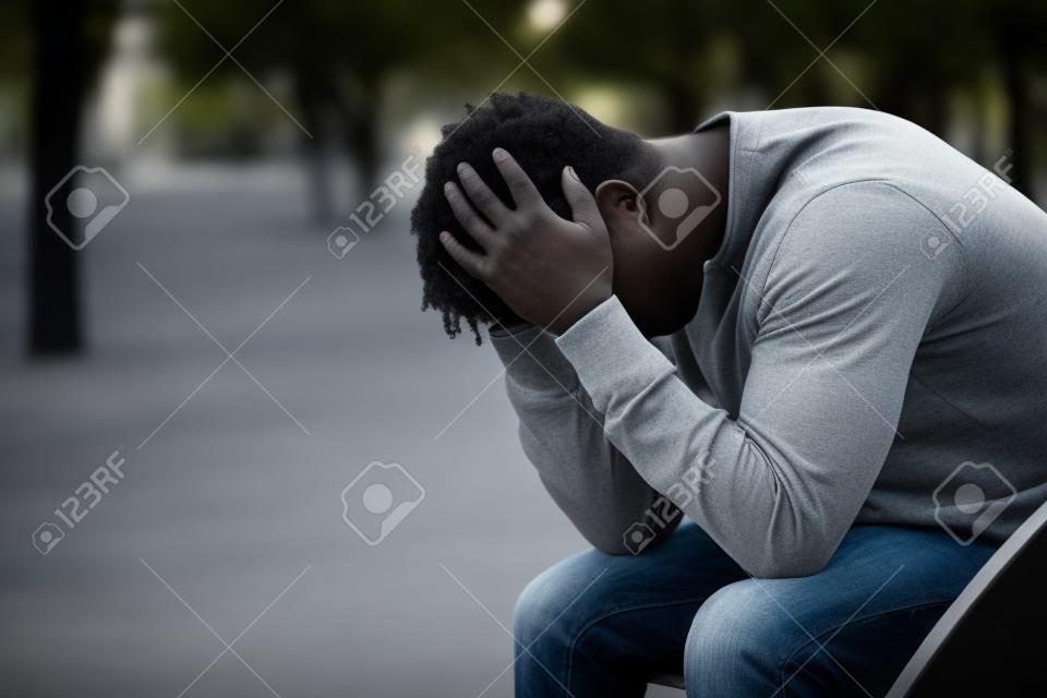 Retrato da vista lateral de um homem preto deprimido triste que senta-se em um banco em um parque